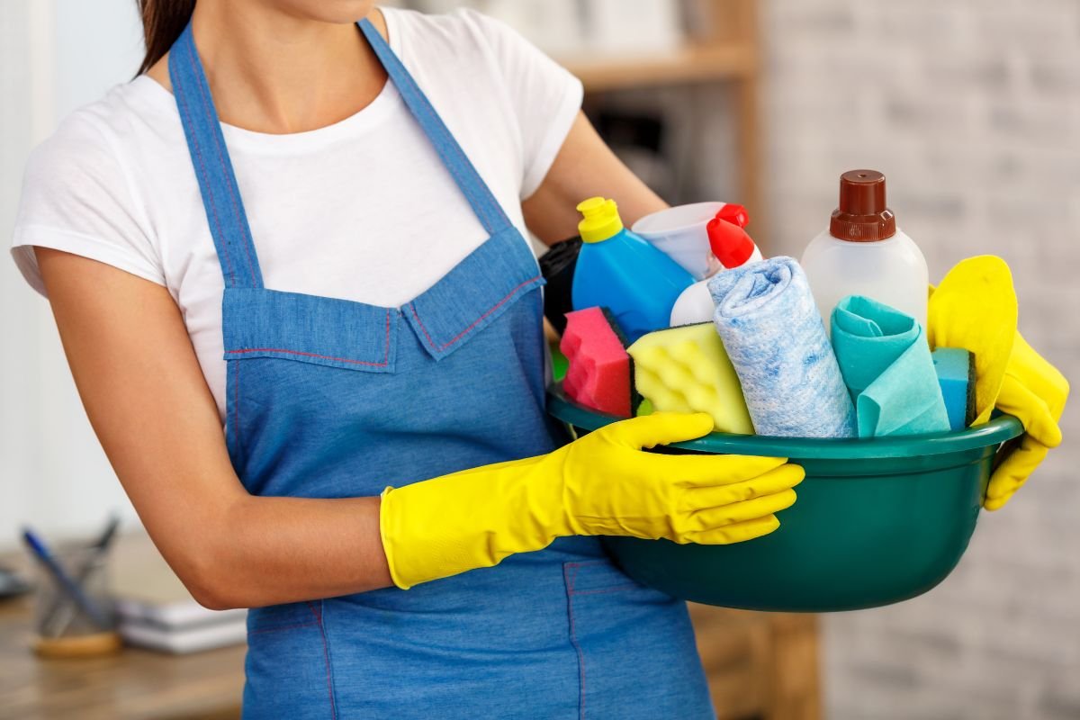 A woman wearing an apron holds a bucket filled with various cleaning supplies, ready to start her cleaning task.