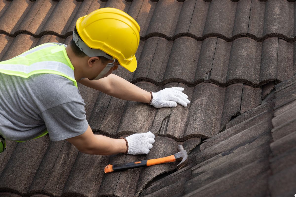 A man wearing a hard hat and safety vest is working on a roof, focused on his task with tools in hand.