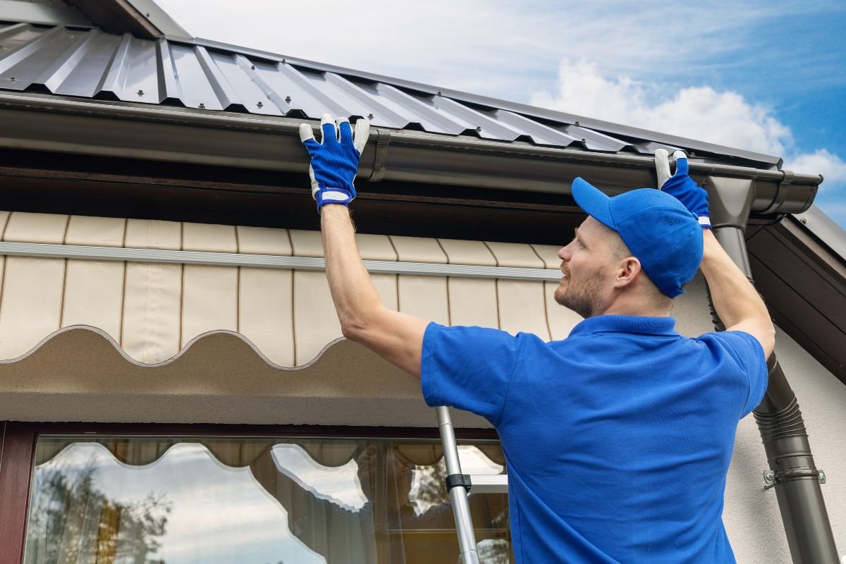 A man wearing a blue shirt and blue gloves repairs a metal roof, focused on his task with tools in hand.