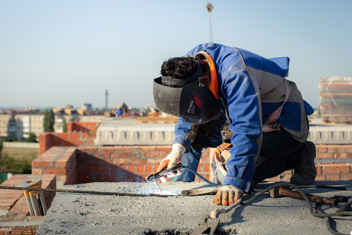 A man wearing a welding helmet works on a roof, using a welding torch to join metal pieces together. 