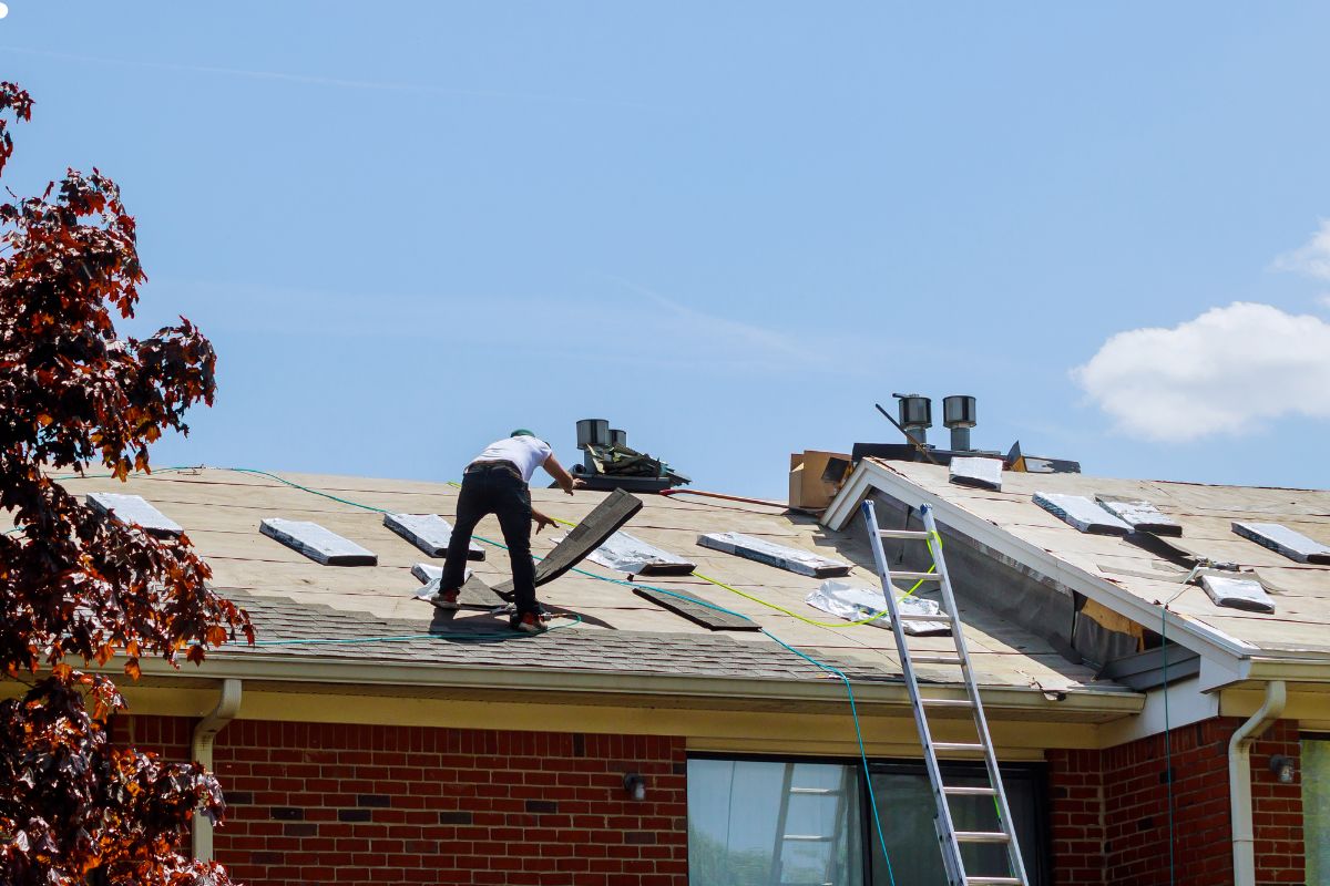 A man is engaged in roofing work, carefully positioning materials on the top of a house under bright sunlight.
